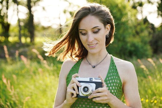 Beautiful young woman wearing green knitted or crocheted top outdoors in nature is taking photographs with old vintage camera