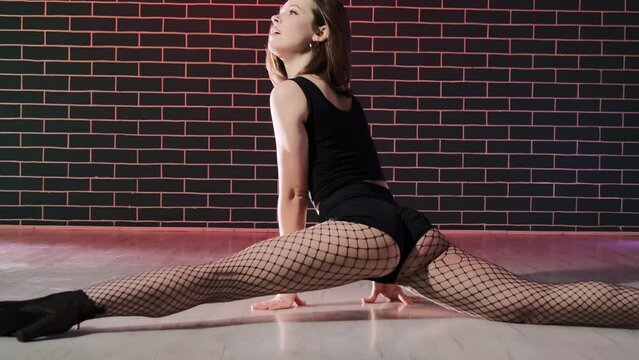Young woman performing erotic dance with split tricks in brick wall studio. Female dancer shows split in black high heels