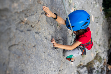 child rock climber in a blue protective helmet overcomes the route in the mountains. children's...