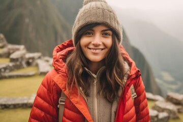 Beautiful young woman in a red jacket and a gray hat is standing on the top of a mountain and smiling.