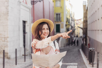 Pretty woman in a straw hat with a paper map walking in a small European street and pointing at...
