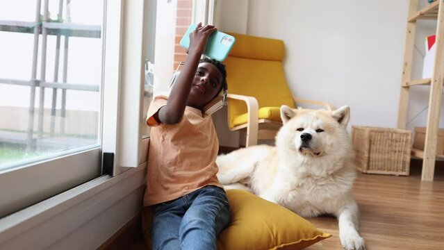 african american boy with headphones and smart phone, takes selfie photos, stands with his dog at home, at the window