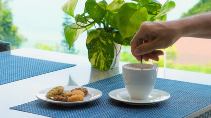 Man stirs coffee in mug with small spoon in sea view cafe on tropical vacation. Beachfront restaurant cafe with breathtaking sea view. Business vacation, holidays in Thailand.