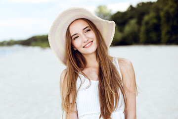 Portrait of a happy smiling woman in free happiness bliss on ocean beach standing with a hat. A female model in a white summer dress enjoying nature during travel holidays vacation outdoors.