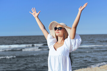 Fototapeta na wymiar Happy blonde woman is on the ocean beach in a white dress, sunglasses and hat, raising hands.
