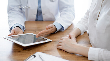 Doctor and patient sitting at the wooden desk in clinic. Female physician's hands pointing into laptop computer monitor, close up. Medicine concept.