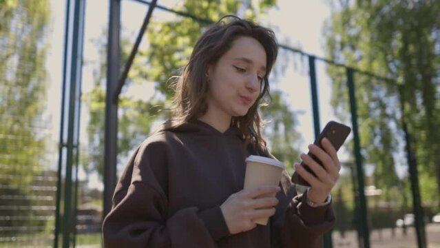 Attractive young girl in a sweatshirt holds a paper cup of coffee in hands and looks into a smartphone on the sports ground. Girl on the street basketball court with a smartphone and a cup of coffee