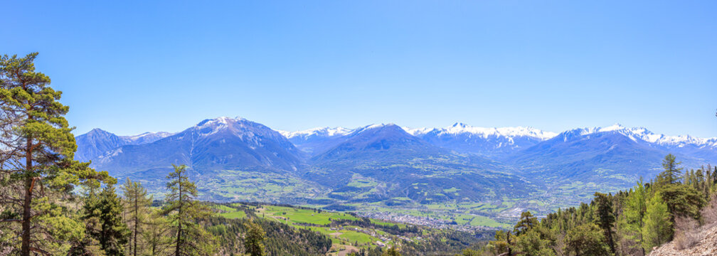 A scenic panoramic view of a mountain valley with a town surrounded by snowy mountain summit under a majestic blue sky