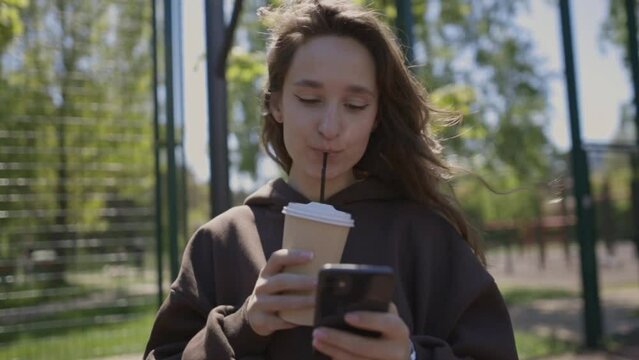 Attractive young girl in a sweatshirt holds a paper cup of coffee in hands and looks into a smartphone on the sports ground. Girl on the street basketball court with a smartphone and a cup of coffee