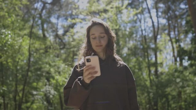An attractive Caucasian girl walks in a city park on a sunny day, and looks into a smartphone. The girl walks in the park and looks at the mobile phone. A girl in the park with a phone in her hands.
