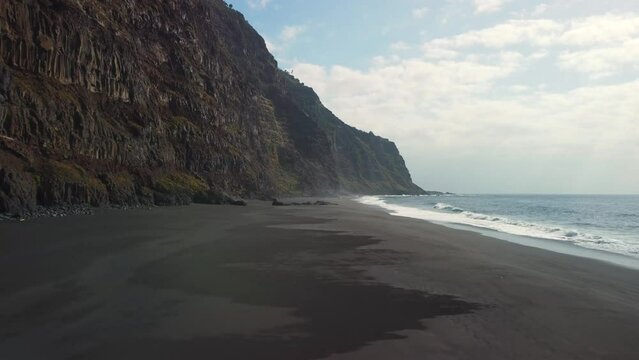 Virgin black sand beach in Tenerife. Giant cliff of volcanic stone. Drone flight over the sand by the sea where the waves break. Paradise beach travel concept