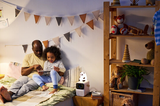 African American dad using digital tablet together with his little son before sleep in bedroom - Powered by Adobe
