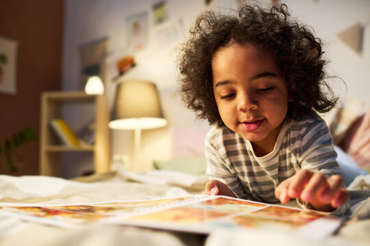 African American boy watching pictures in book before sleep while lying on bed in bedroom