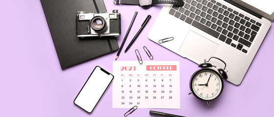 Calendar for October 2023, alarm clock, laptop, mobile phone, photo camera and office supplies on lilac background