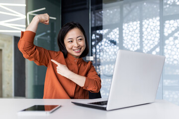 Fototapeta na wymiar A young Asian woman working in the office using a laptop, talking on a video call and showing a power pose to the camera, pointing a finger at her hand. Brag about your success and achievements.