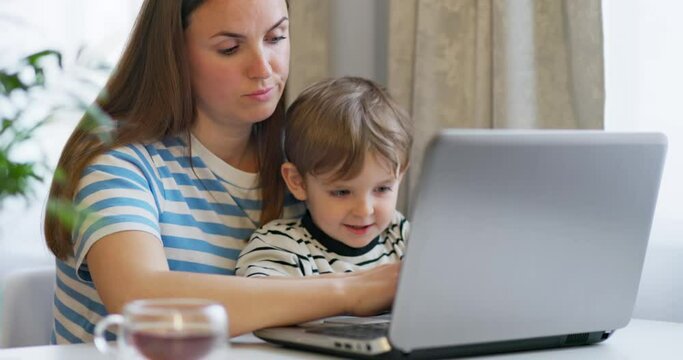 Mother working on laptop from home remotely with little son in her arms. 