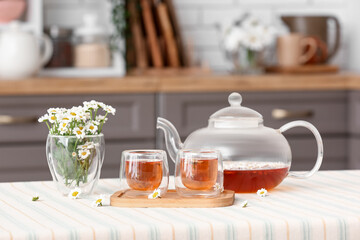 Wooden tray with teapot, cups of natural chamomile tea and flowers on table in kitchen