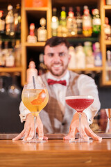 Young mixologist serving two colorful cocktails to his customers with a smile. Concept: Drinks, fun, nightlife