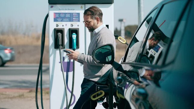 Video of elegant man using mobile phone while charging electro car at the electric gas station
