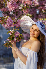 A beautiful girl in a white short airy dress and a white wide-brimmed hat on vacation near cherry blossoms