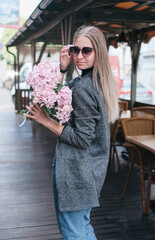 Beautiful caucasian girl with blond hair in a gray coat and blue jeans with a bouquet of pink hydrangeas