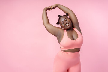 Smiling African American sporty woman wearing sportswear stretching, training, doing exercises isolated on pink background. Happy beautiful plus size model posing for pictures. Body positive concept
