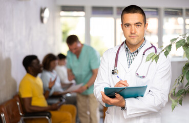 Portrait of focused man, professional doctor wearing white coat standing in lobby of clinic, filling out medical form at clipboard - obrazy, fototapety, plakaty