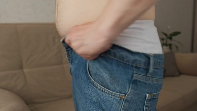 A pot-bellied man is trying to pull denim pants on an obese waist in a close-up room, the side view is unrecognizable. The concept of malnutrition and overeating
