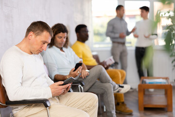 Serious man browsing websites on smartphone while sitting in lobby of job center or employment...