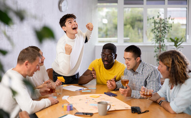Fototapeta premium Excited happy young guy having fun with multiethnic group of male friends, playing board game at table
