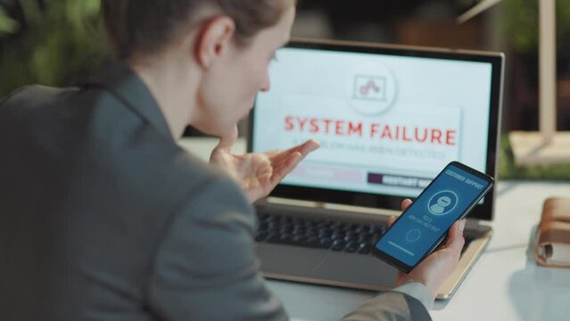 Seen from behind modern business woman with laptop and smartphone having pc fail and talking with voice bot in modern office.