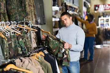 Positive guy choosing camouflage uniform in military shop