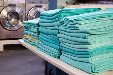 Stack of folded green surgical cloths in an industrial laundry and washing machines behind.