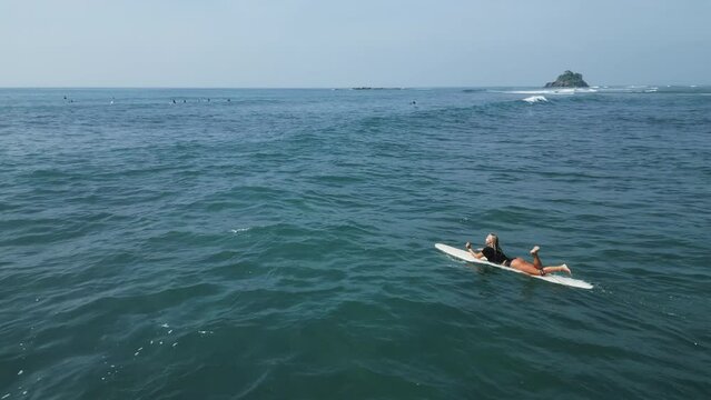 Young female surfer rowing on surfboard in crystal ocean water at scenic tropical beach on sunny day aerial view. Surfing girl enjoying sport paddling and surfing on summer vacation drone shot