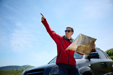 A man leans against a car, holding a map in his hands, as he looks for directions. The traveler...