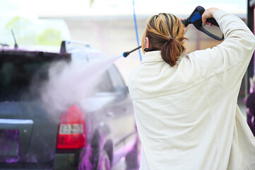 Fototapeta na wymiar A young woman wearing glasses and casual attire cleans her car using a high-pressure washer on a bright sunny day after driving through a dirty terrain.