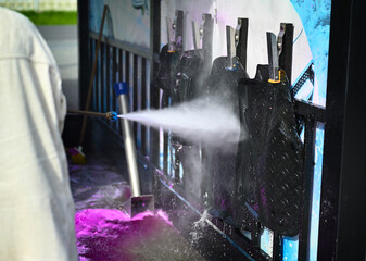 A young woman wearing glasses and casual attire cleans her car using a high-pressure washer on a bright sunny day after driving through a dirty terrain.