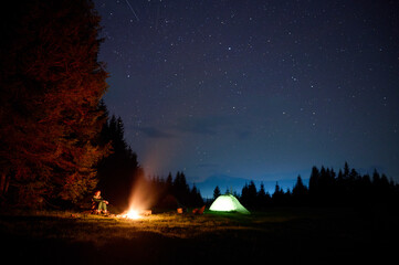A young female traveler sits by the campfire near her tent, enjoying a moment of relaxation beneath the beautifully starry sky. As the crackling flames provide warmth and comfort