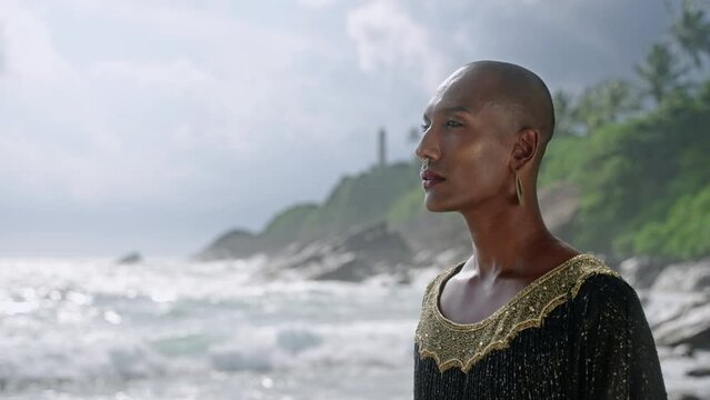 LGBTQIA black person in luxury dress on stunning sea shore closeup. Gender fluid ethnic stylish model wearing jewellery dressed in posh gown in tropical location against dramatic sky portrait