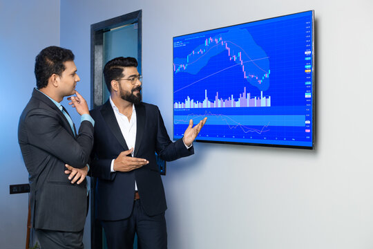 Indian businessmen colleagues trading and discussing stocks online in office. Stock brokers looking at indexes graphs on big computer screen.