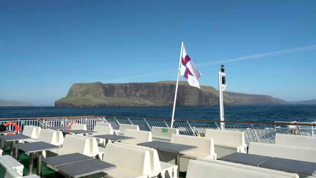 Waving Faroese Flag on Smyrill ferry with Sandoy Island in background - Blue sky and sunlight in Faroes Islands