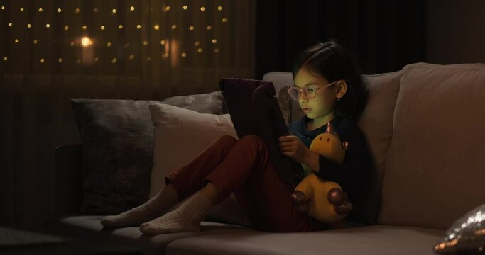Cute girl with digital tablet sitting on sofa at home at night