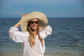 A happy blond woman with sunglasses is standing by the sea. She is touching her straw hat. A girl with long hair and a white shirt spends her vacation on the beach. Holidays to other countries