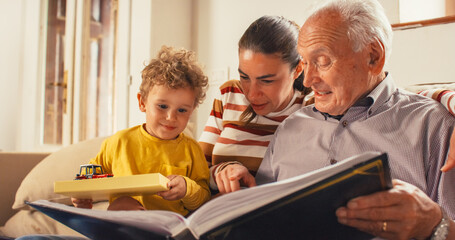 Portrait of a Small Family of Mother, Son, and Grandfather Watching a Photo Album in the Living...