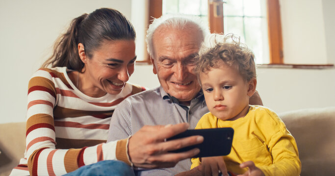 Happy Small Family Gathered Together, Sitting on a Sofa in the Living Room and Using a Smartphone. Mother Showing Photos to her Kid on her Mobile Phone in the presence of the Grandfather