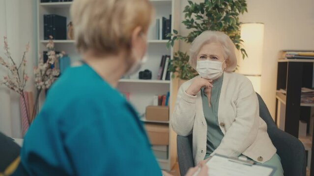 Doctor and senior patient in face masks talking in room, home visiting service