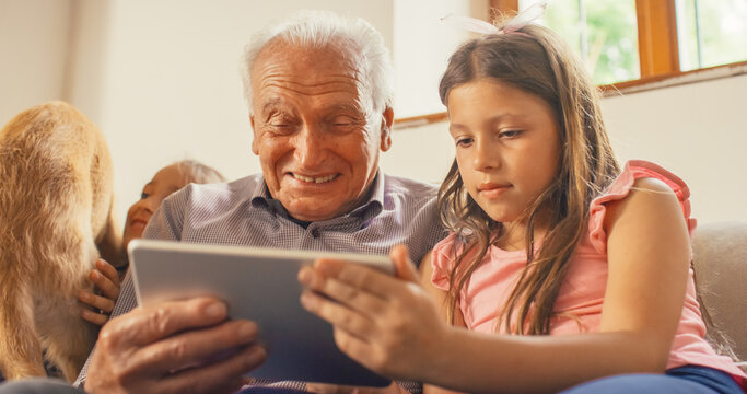 Happy and Laughing Senior Man Enjoying the Company of his Grandchildren and Family Dog While Using Digital Tablet in the Living Room. Girls Spending Time with their Grandfather who is Telling Jokes