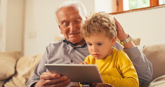 Portrait of a Senior Man Using a Tablet to Read a Children's Book to his Little Grandson While Sitting on a Sofa. Cute Child Listening Carefully to a Story that his Grandfather is Telling