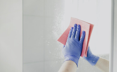 Cropped view of housekeeper woman hand in blue rubber glove polishing, cleaning, swiping mirror with cloth rag, spraying cleaner liquid while cleaning bathroom at home. Housekeeping, cleaning service
