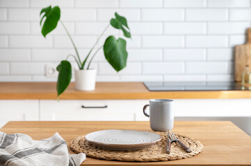 Wooden table top with plate and cup for breakfast against blurred white kitchen with cutting board and monstera plant in scandinavian style